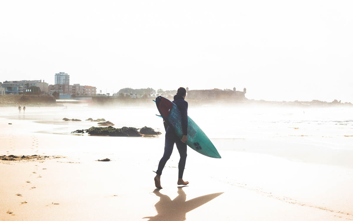 man walking on beach in matosinhos with surfboard under arm in porto portugal